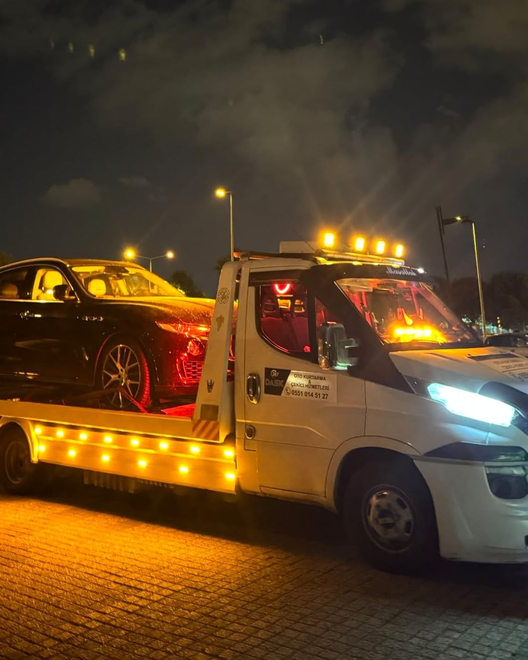 Gece tepe lambaları yanan Dask Oto Kurtarma çekicisi, kasasında siyah otomobil